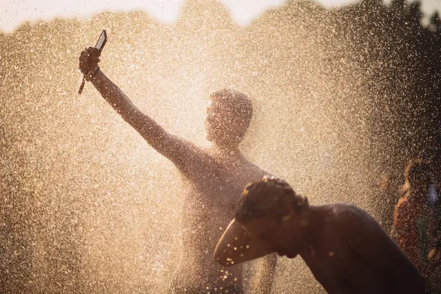 Twee mensen koelen zich af onder een opspattende waterstraal in fel tegenlicht op een warme zomerdag