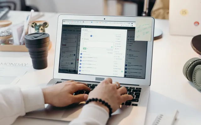 Iemand typt op een laptop aan een licht bureau, met een koffiebeker en een koptelefoon ernaast